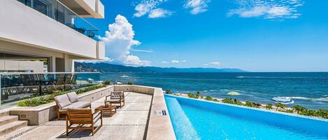 A rooftop pool with a view of the sea, outdoor seating, and a modern building.