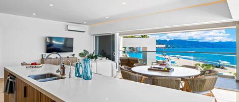 A modern kitchen with a large island, a dining area with wicker furniture, and a view of a beach and ocean.