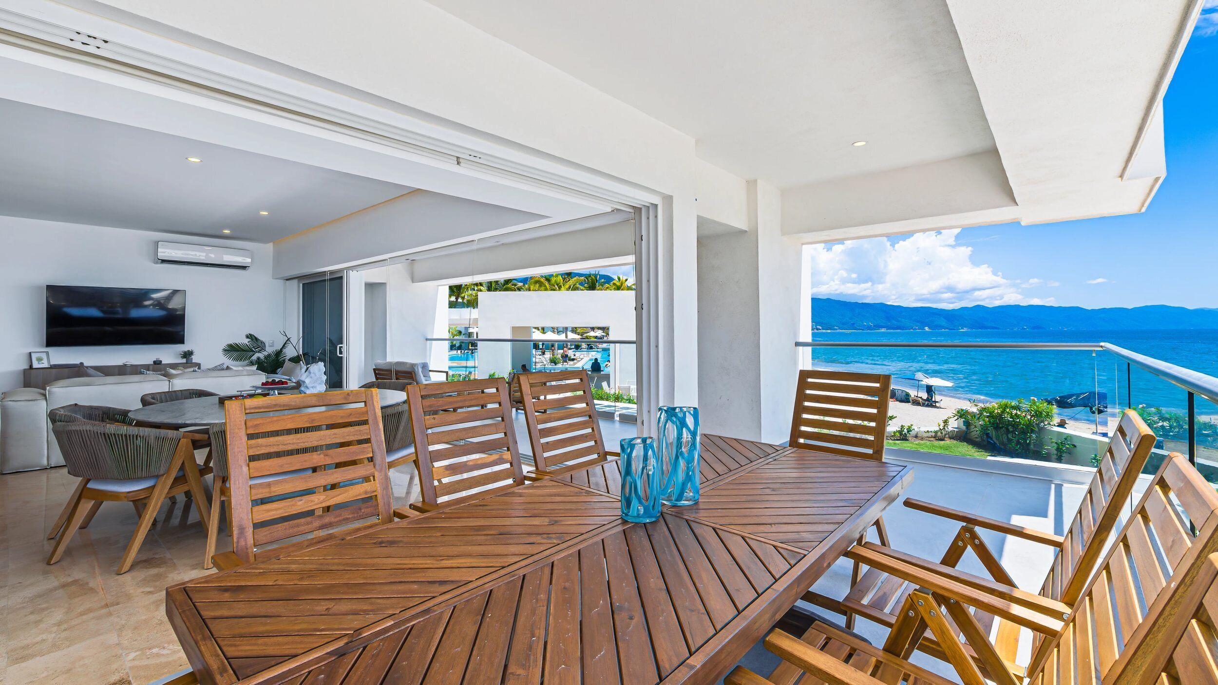 Een houten eettafel met zes stoelen, een aan de muur bevestigde televisie en een balkon met uitzicht op zee.
