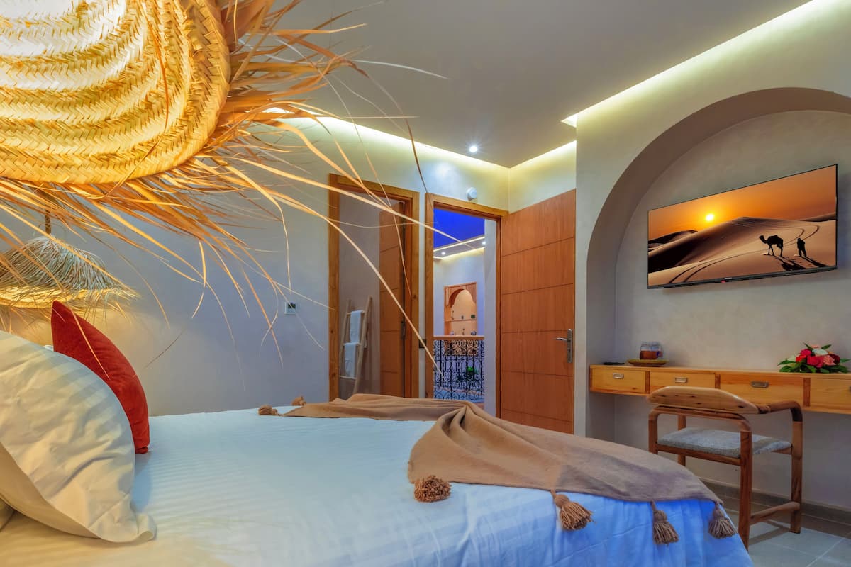 A bedroom with a bed, a desk, and a chair. There is a painting on the wall and a door leading to another room.