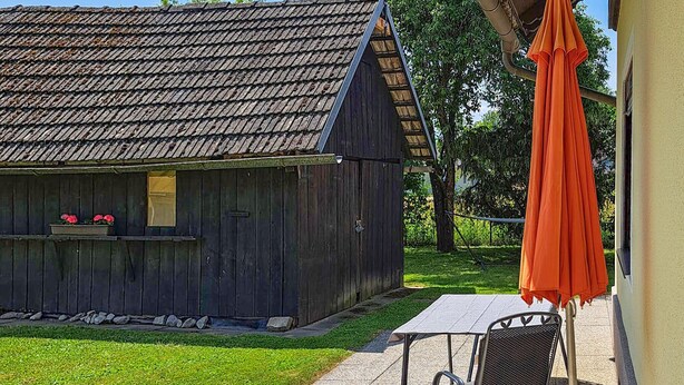 A wooden building with a sloped roof, a small outdoor seating area with a table and chairs, and a hanging orange umbrella. - Lovely apartment in Strau with WiFi (Strau)