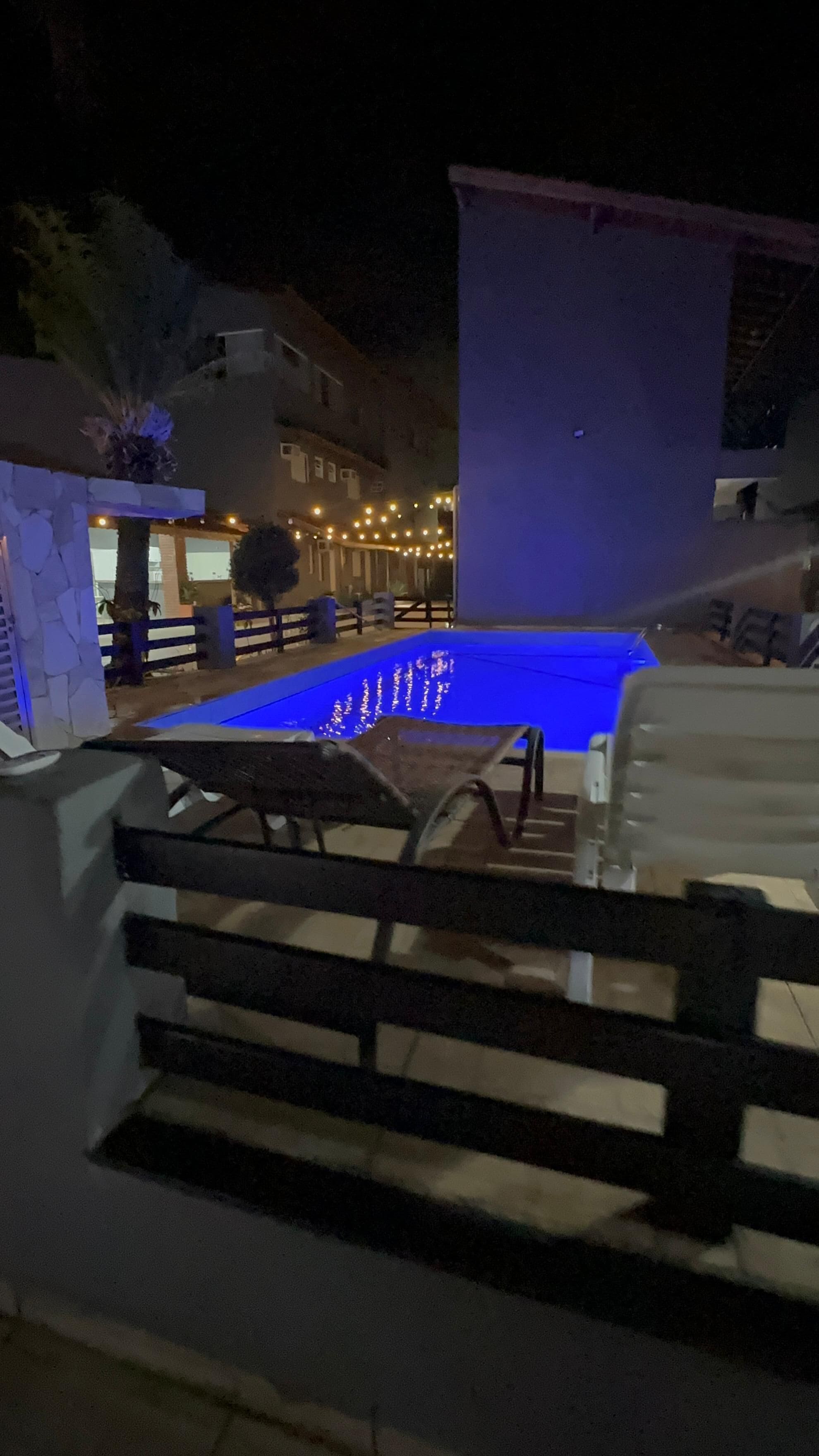 Una piscina con un deck iluminado en azul, una silla de descanso y un muro de piedra.