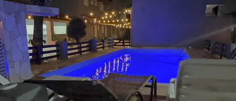 Una piscina con un deck iluminado en azul, una silla de descanso y un muro de piedra.