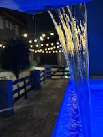 Ambiente esterno notturno con giochi d'acqua, sentiero in pietra e luci a catena.