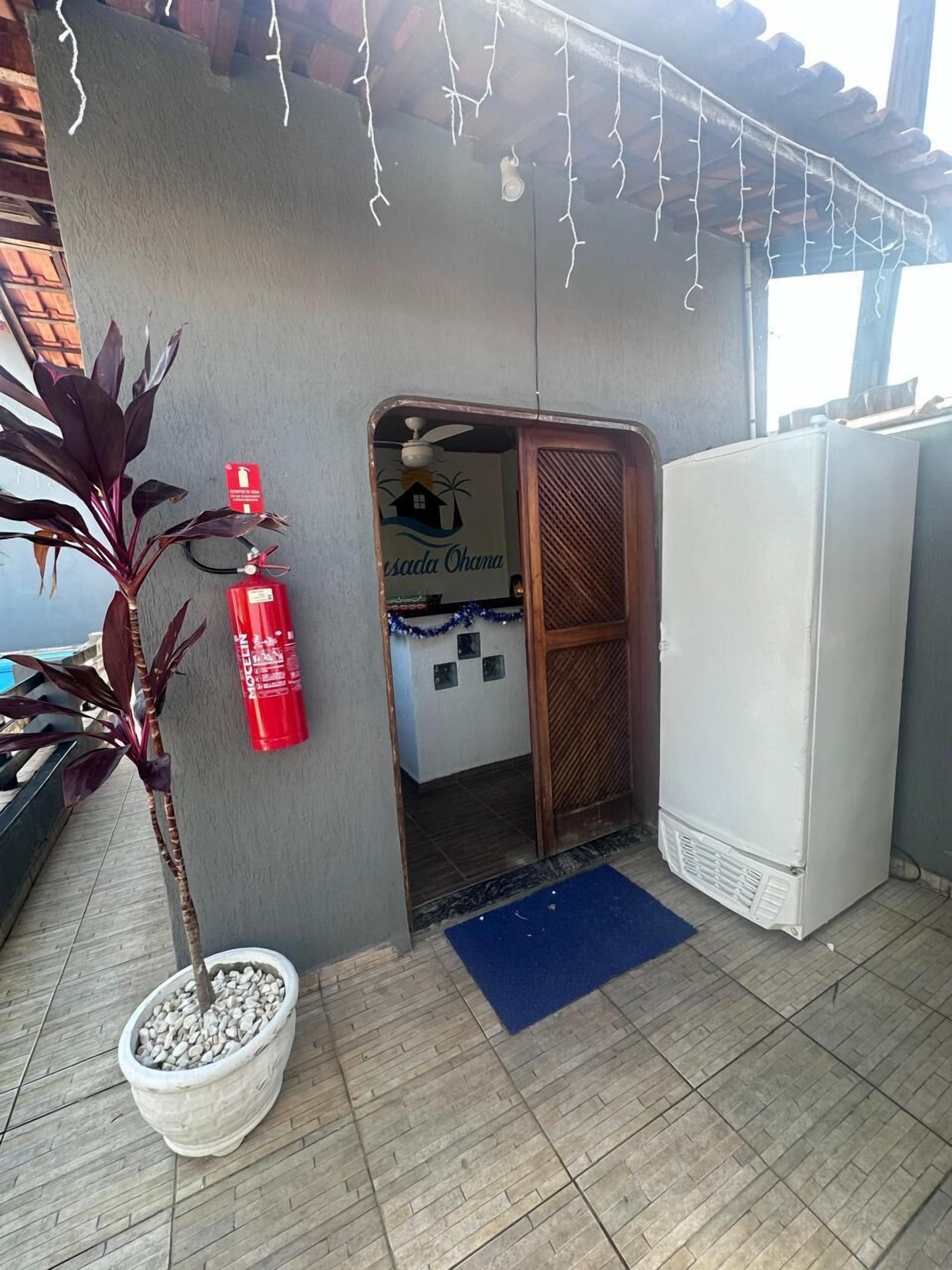 Una habitación pequeña con una puerta que da a un balcón, un refrigerador blanco, una planta en maceta y un extintor.
