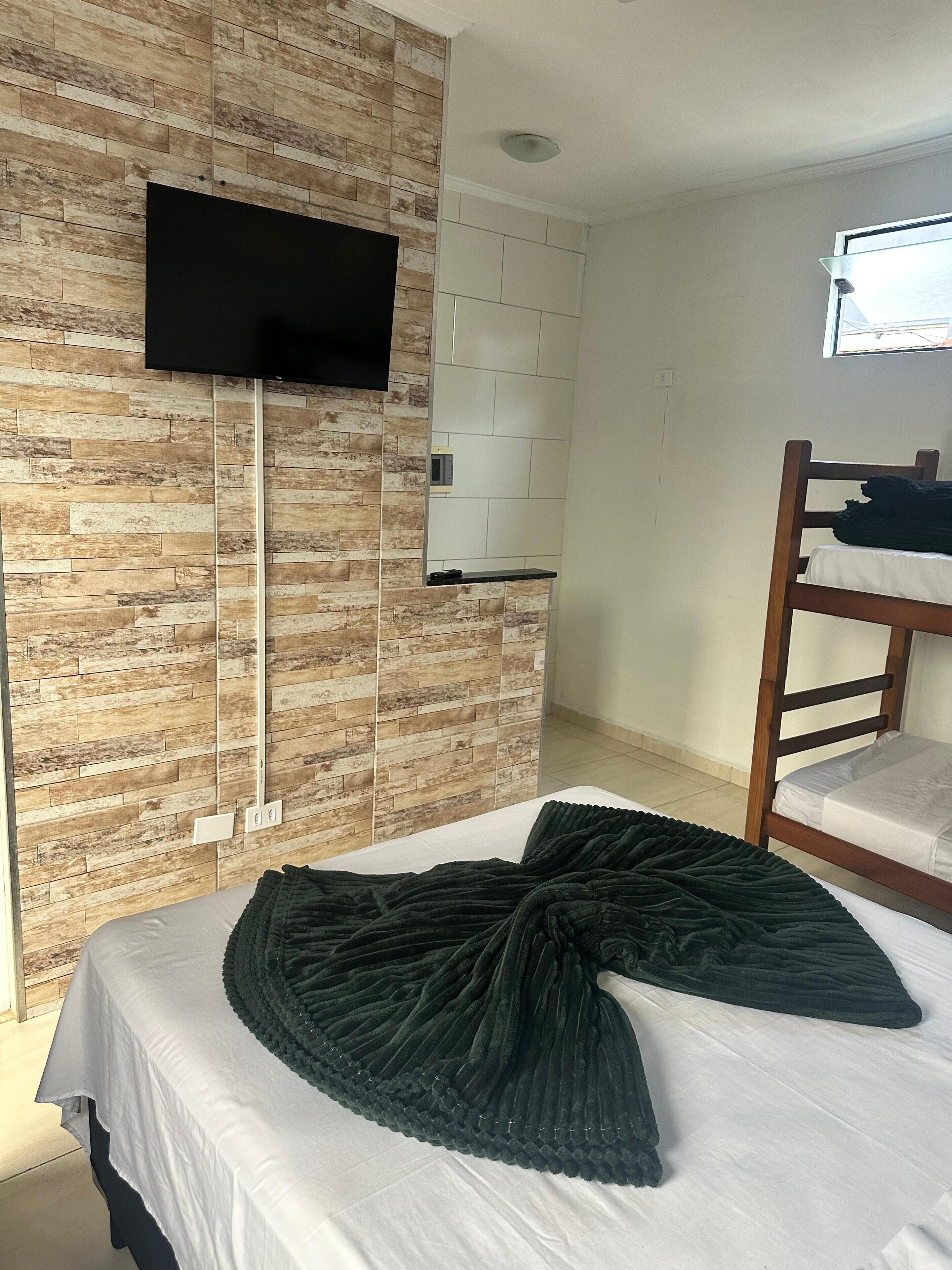A bedroom with a brick-patterned wall, a flat-screen TV mounted above a shelf, a bunk bed, and a large, dark green, textured blanket on the bed.