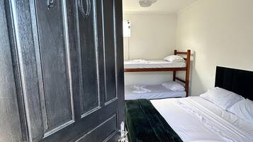Quarto com porta de madeira escura, cama com roupa de cama branca, manta verde, ventilador de teto e uma cama beliche ao fundo.