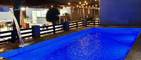 Una piscina con fuente de agua, rodeada de sillas de descanso y un edificio con iluminación exterior.