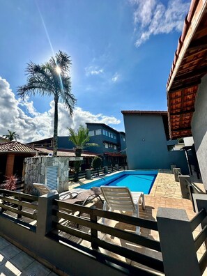 A swimming pool with lounge chairs, a stone wall, and a palm tree. - pousada ohana (Bertioga)