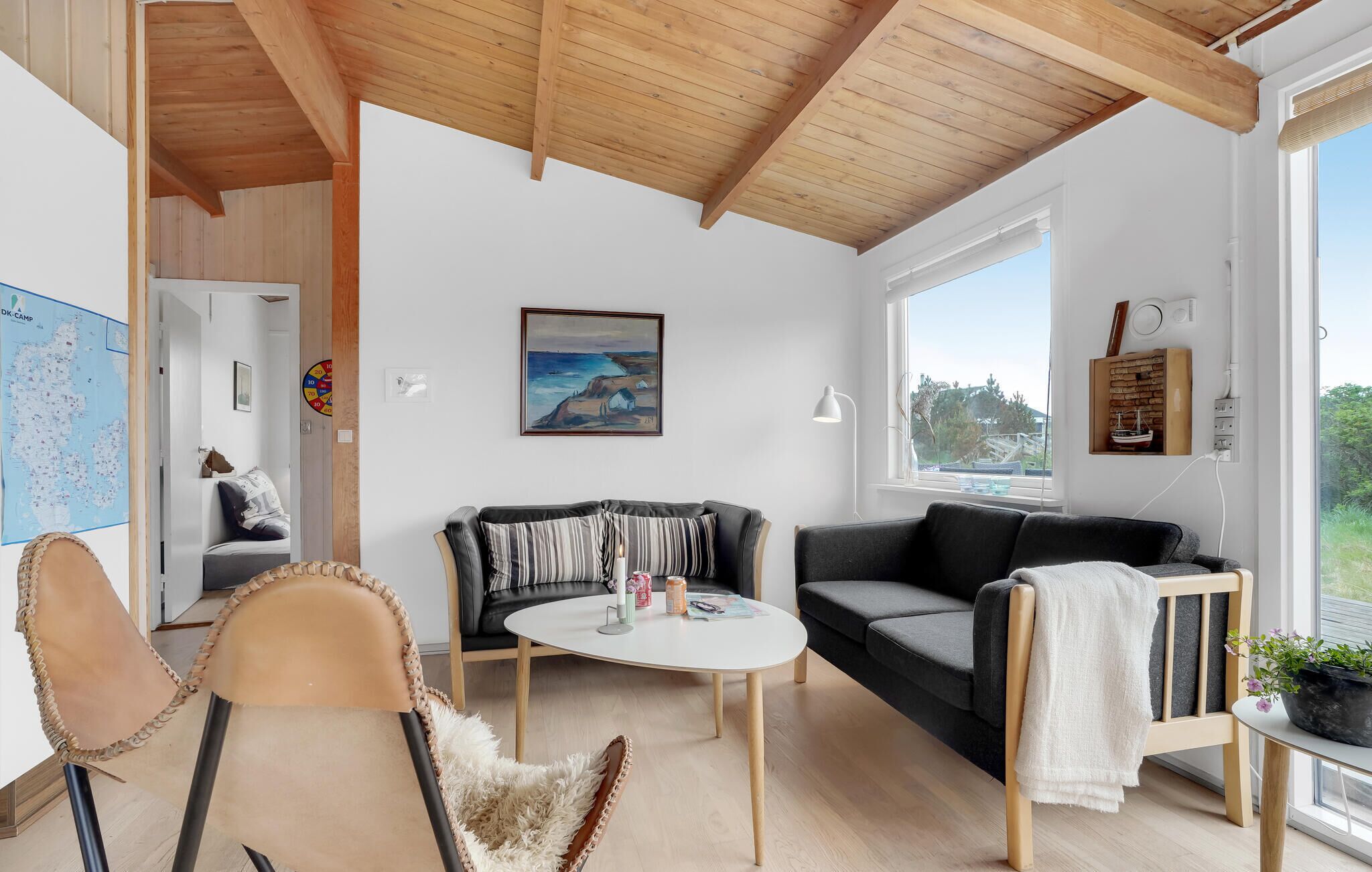 Ein gemütliches Wohnzimmer mit Holzbalkendecke, einem schwarzen Sofa, einem weißen Couchtisch und Blick ins Freie.