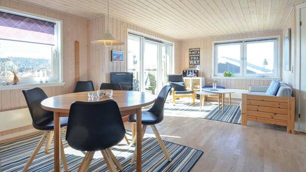 Ein gemütlicher, gut beleuchteter Raum mit Holzbalkendecke, großen Fenstern, einem runden Esstisch mit Stühlen, einer Couch und einem kleinen Tisch mit Lampe.