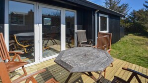 Eine Holzterrasse mit Tisch und Stühlen, eine Schiebetür zu einem Sitzbereich im Freien und ein Rasengrundstück.