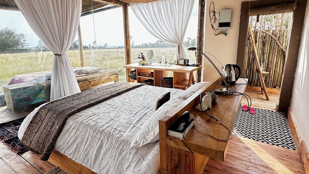 A rustic bedroom with a wooden bed, a desk with a lamp, and a view of the outdoors. - Sironka Cabin Getaway in Ole Lekurruki, Naromoru. (Mureru Farm)