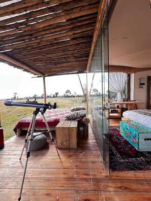 A bedroom with a large window, wooden flooring, a telescope, and a bed with a colorful blanket.