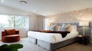 A modern bedroom with a large bed, a red armchair, a bedside table with a lamp, and a window with curtains.