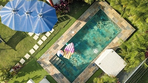 An aerial view of a swimming pool with a tiled deck, surrounded by grass and palm trees.