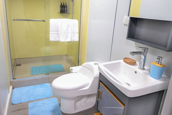 A bathroom with a glass shower enclosure, a white toilet, a sink with a soap dish, and a blue mat on the floor.
