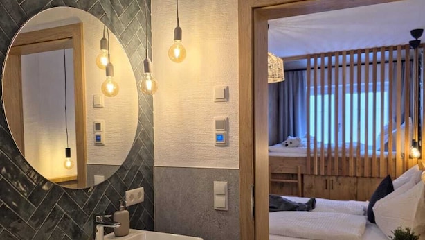 Un baño moderno con un espejo grande, un lavamanos y un mueble de tocador de madera. Hay una cama con cabecera de madera y una ventana con persianas.