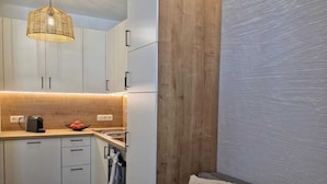 Cocina compacta con armarios blancos, pared de acento de madera, lámpara colgante y sofá gris con cojines.