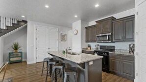 Une cuisine moderne dotée d’un îlot central, de tabourets de bar, de meubles de couleur sombre, d’appareils électroménagers en acier inoxydable et d’un escalier menant à un autre niveau.