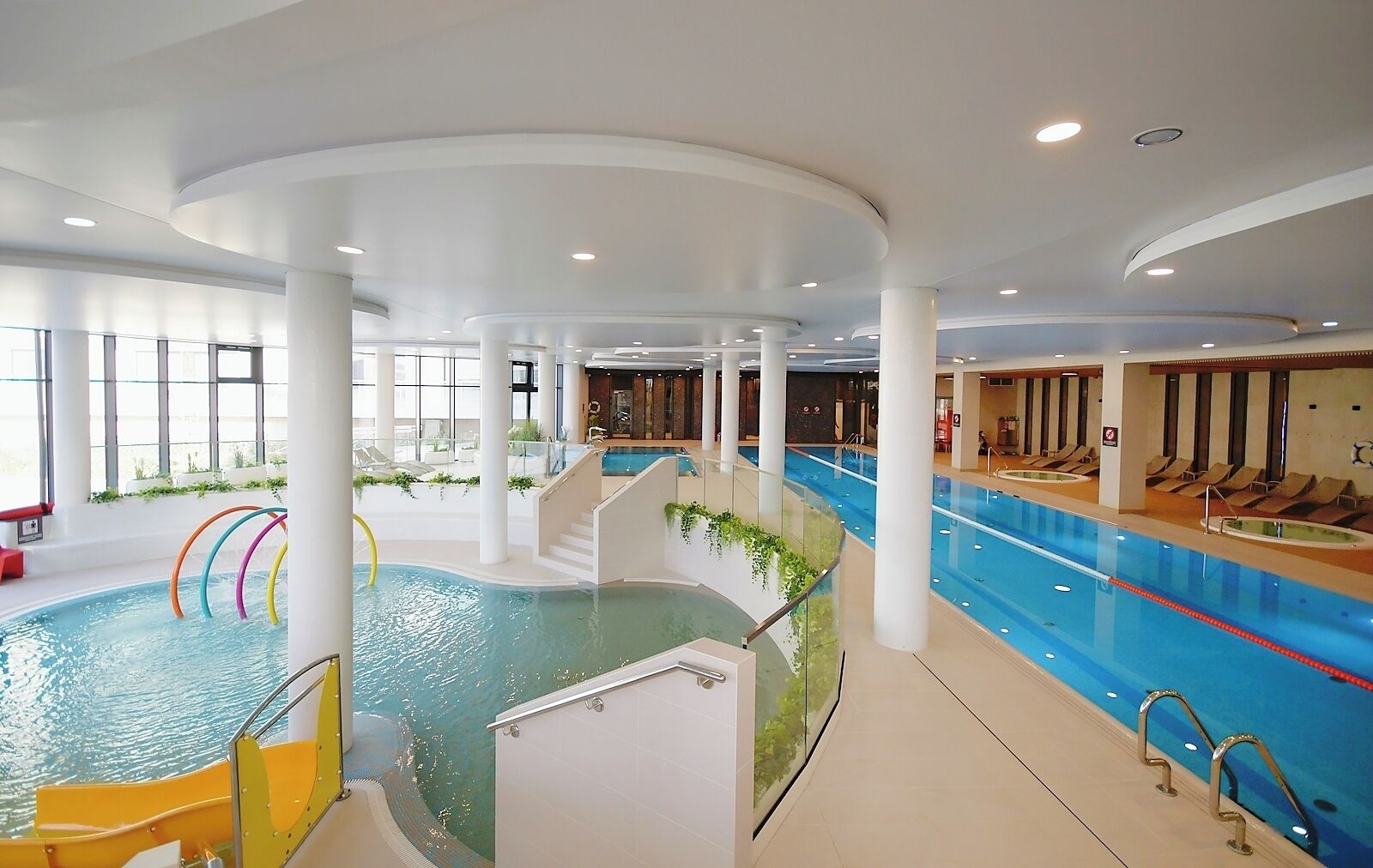 An indoor swimming pool with multiple lanes, a changing room area, and a seating area with sun loungers.