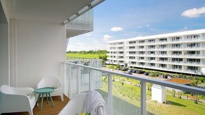 A balcony with white furniture, a view of a green park, and a multi-story building in the background.