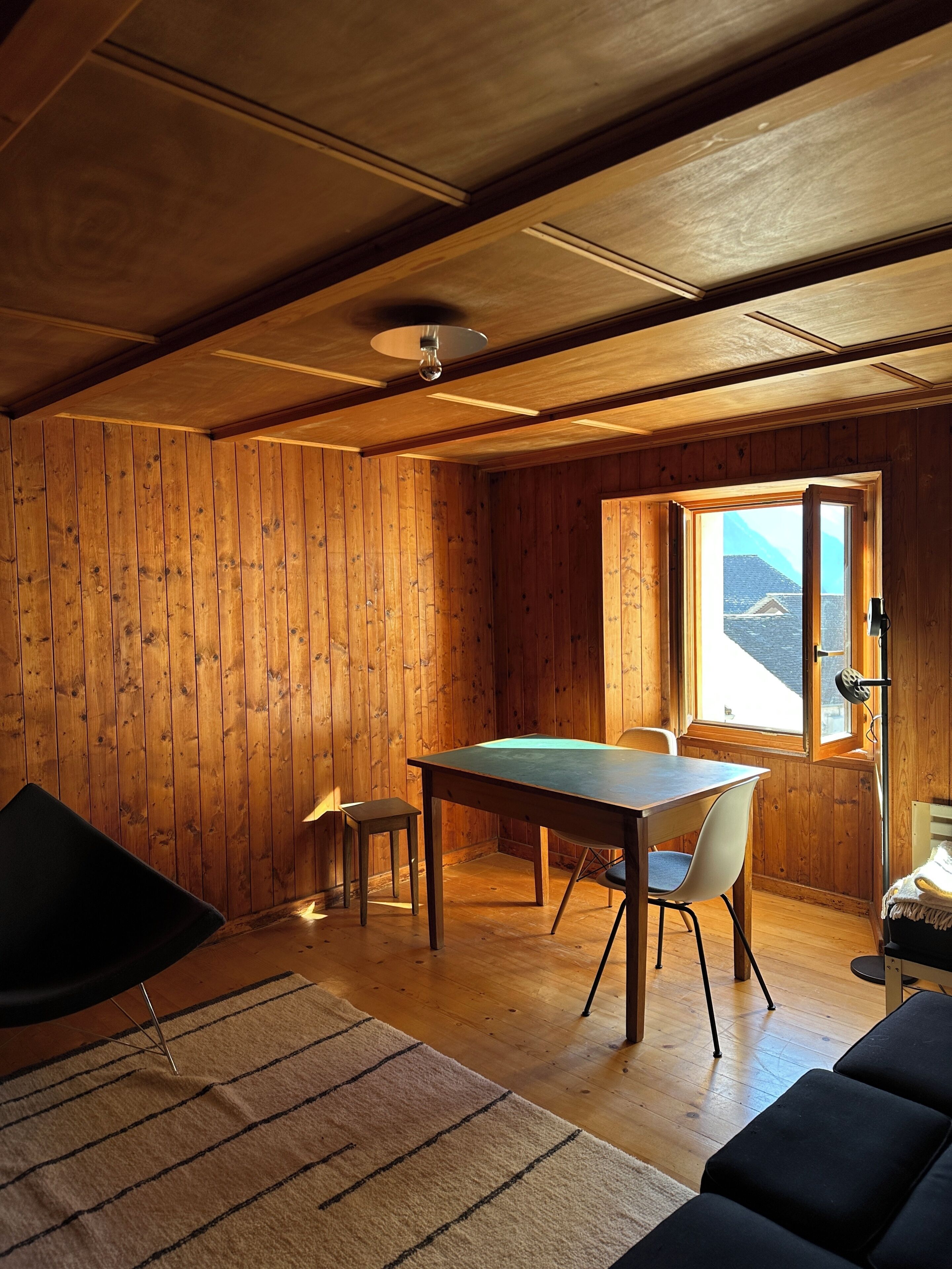 A room with wooden walls, a dining table with chairs, a small stool, a sofa, and a window with a view.