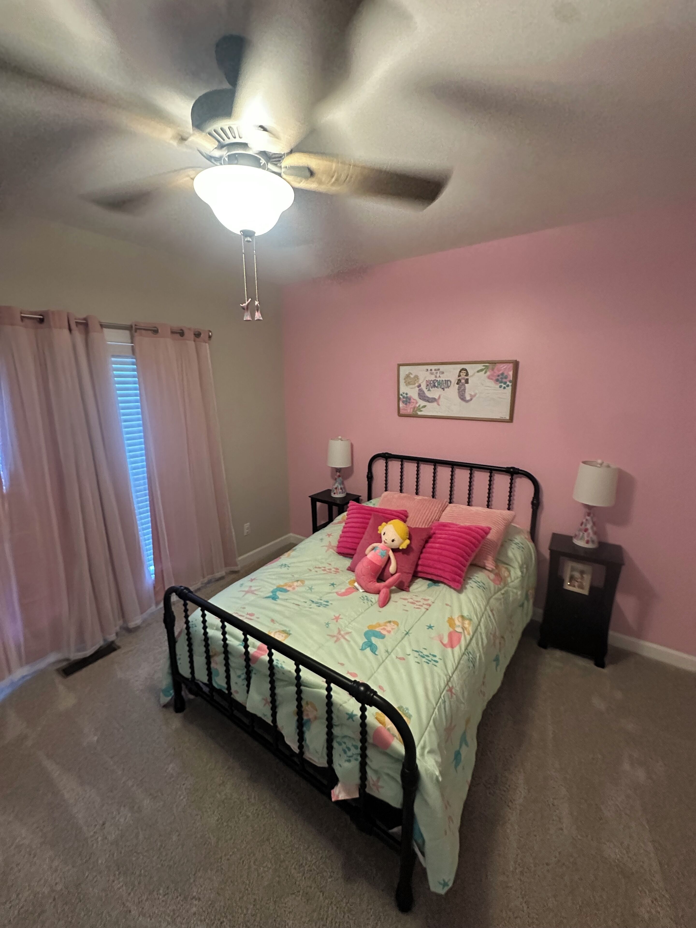 Une chambre à coucher avec un lit, un ventilateur de plafond, une fenêtre avec des rideaux et un élément décoratif mural.