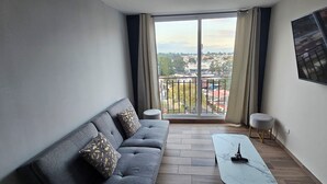 Un salon moderne avec un canapé gris, une petite table basse blanche et un balcon offrant une vue sur le paysage urbain.