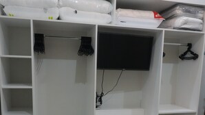 Un dormitorio con una cama, un armario blanco con varios estantes y cajones, y un televisor montado en la pared.