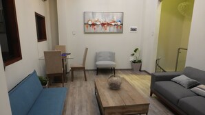Una sala de estar con un sofá azul, un sillón gris y un sofá gris. Hay una mesa de centro de madera con un jarrón decorativo. Un cuadro cuelga de la pared sobre el sillón.