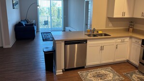 A modern kitchen with a dishwasher, sink, and white cabinets. There is a dining area with a blue sofa and a view of the outdoors through large windows. - Chic 2-bedroom apartment with AC, WiFi in wonderful Los Angeles (Los Angeles)