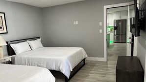 A hotel room with two beds, a bedside table, a TV mounted on the wall, and a view into the kitchen area.