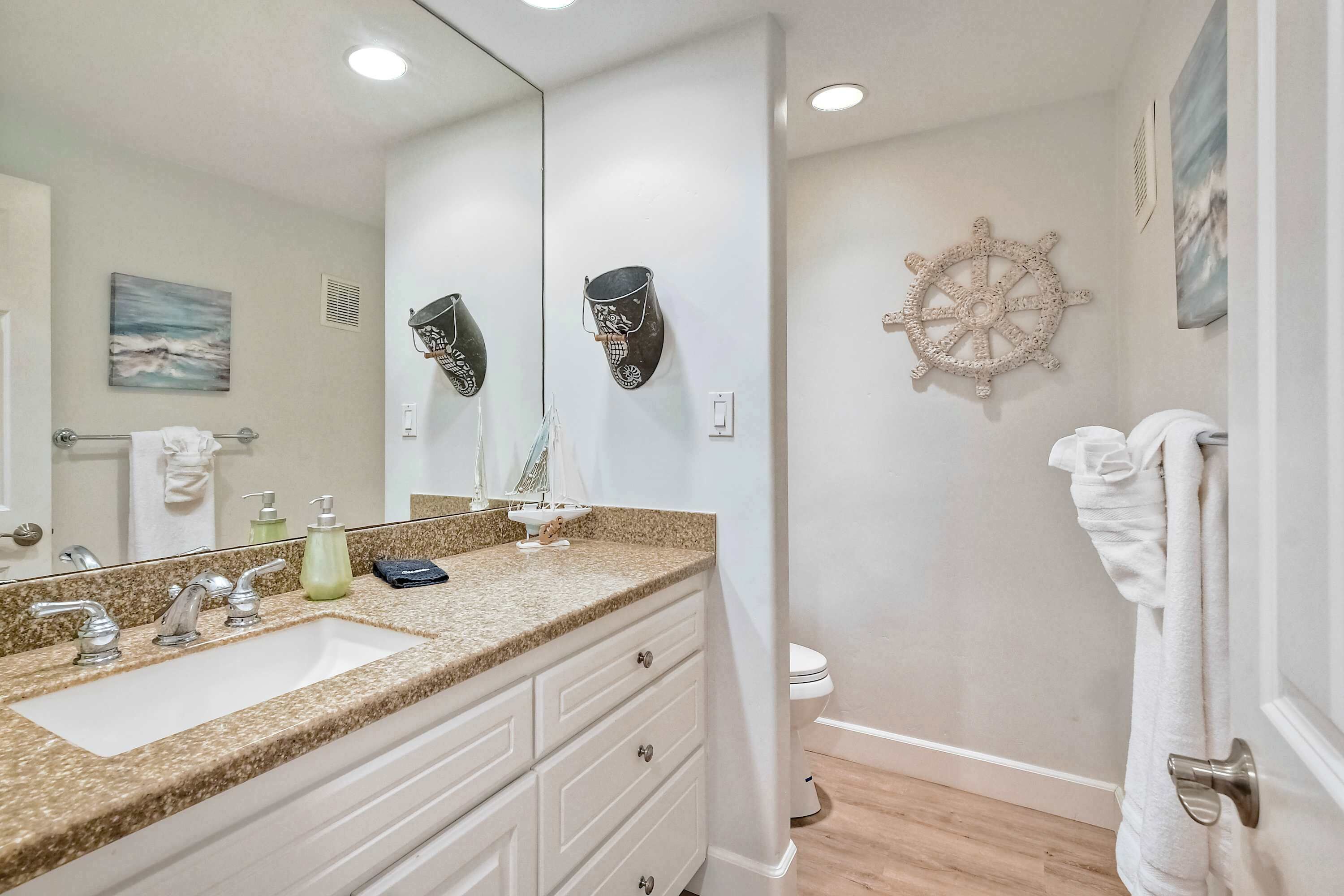 Une salle de bain avec un lavabo sur un comptoir en granit, des meubles blancs, un grand miroir et une décoration en forme de roue de navire au mur.