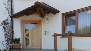 A building entrance with a wooden door, a potted plant, a pathway, and a window with a curtain. - Sommerhof Rauber – Studio 'A 03' with Lake View, Shared Garden and Wi-Fi (Immenstaad am Bodensee)