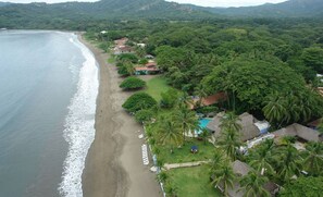 An aerial view of a coastal resort with a beach, palm trees, and thatched-roof buildings. - Beautiful Casa Azul (Potrero)