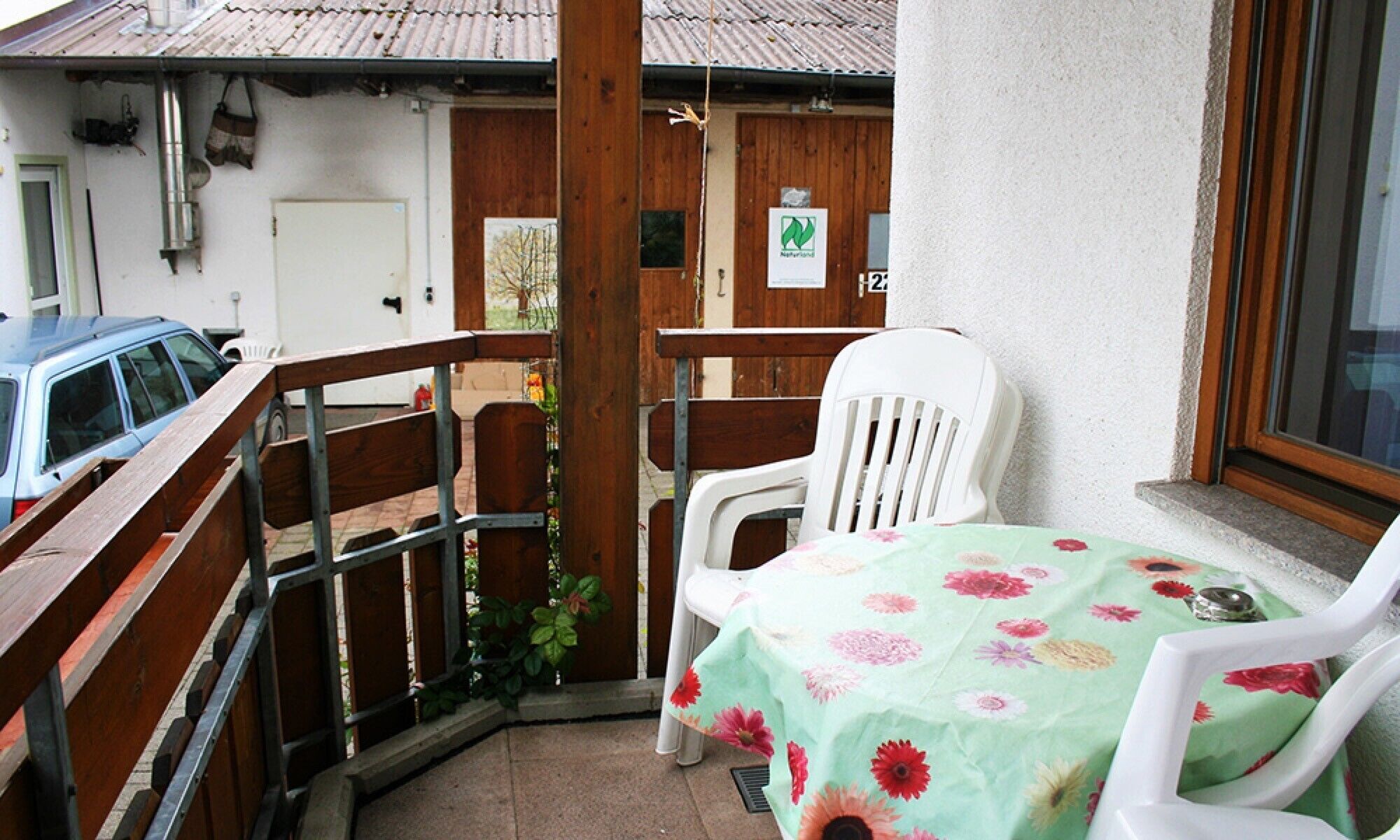 A balcony with a wooden railing, a white plastic chair, a table with a floral tablecloth, and a parked car.