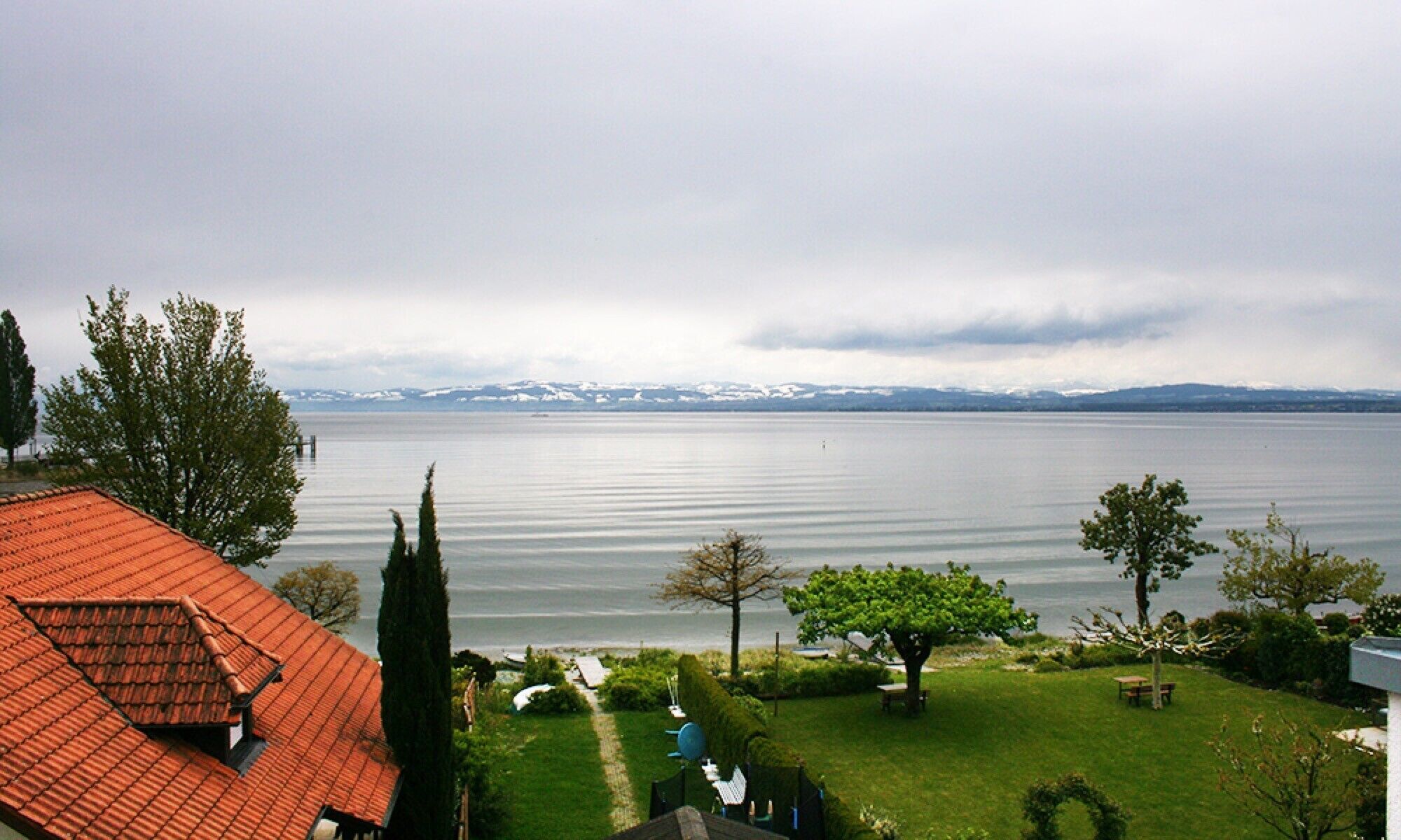 Ein Haus mit rotem Dach, einer Rasenfläche und Blick auf ein großes Gewässer mit Bergen im Hintergrund.