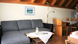 Una sala acogedora con un sofá gris, una mesa de comedor de madera y sillas. Un cuadro enmarco cuelga en la pared, y se observa una lámpara de pie.