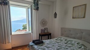 A bedroom with a large bed, a view of the sea, a small table with a fan, and a picture on the wall.