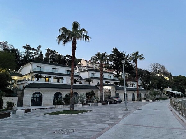 Un edificio de varios pisos con un camino pavimentado, palmeras y un coche estacionado al frente.