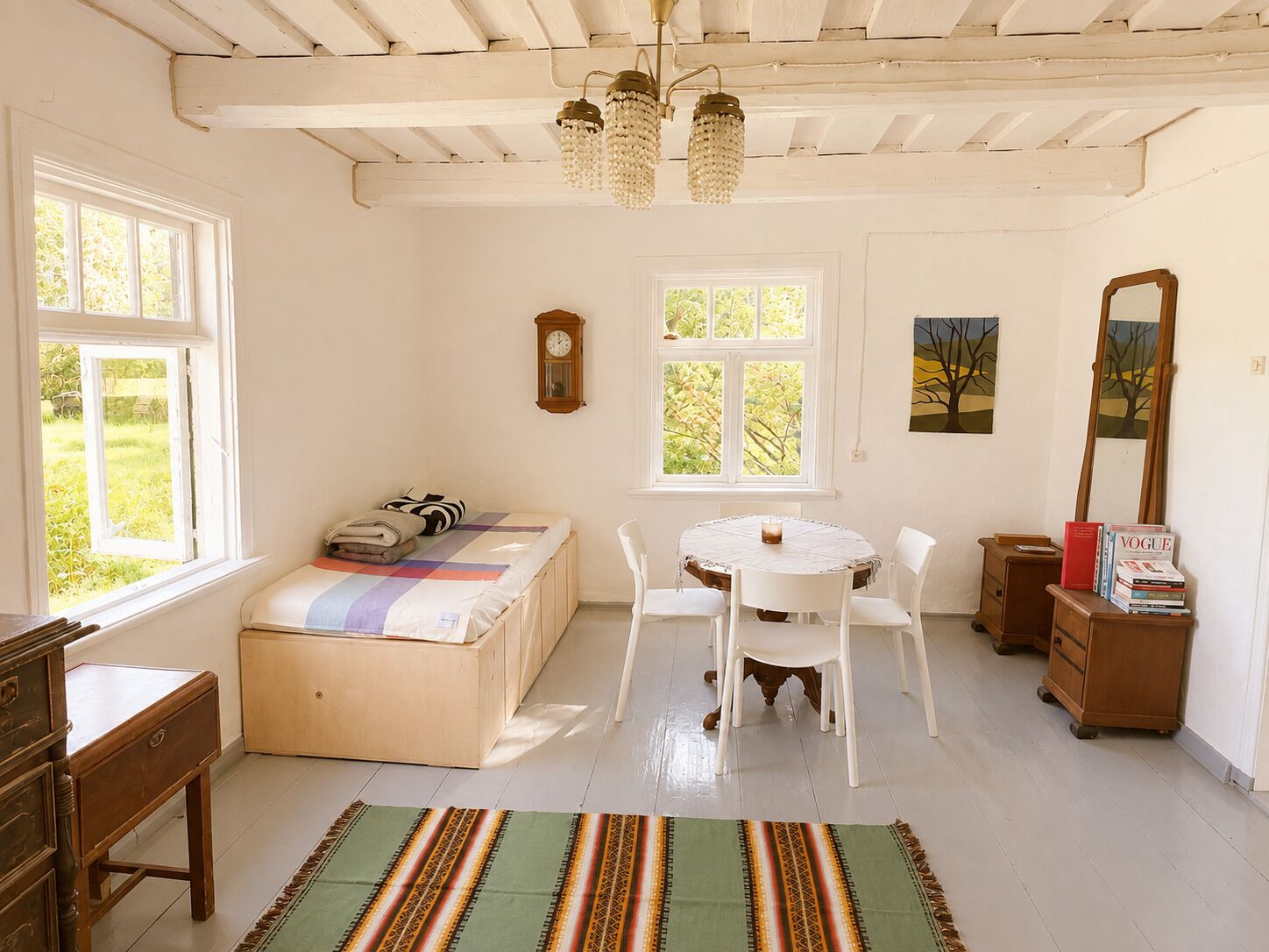 A room with a bed, a round table with chairs, a wooden dresser, a mirror, and a window with a view of greenery.