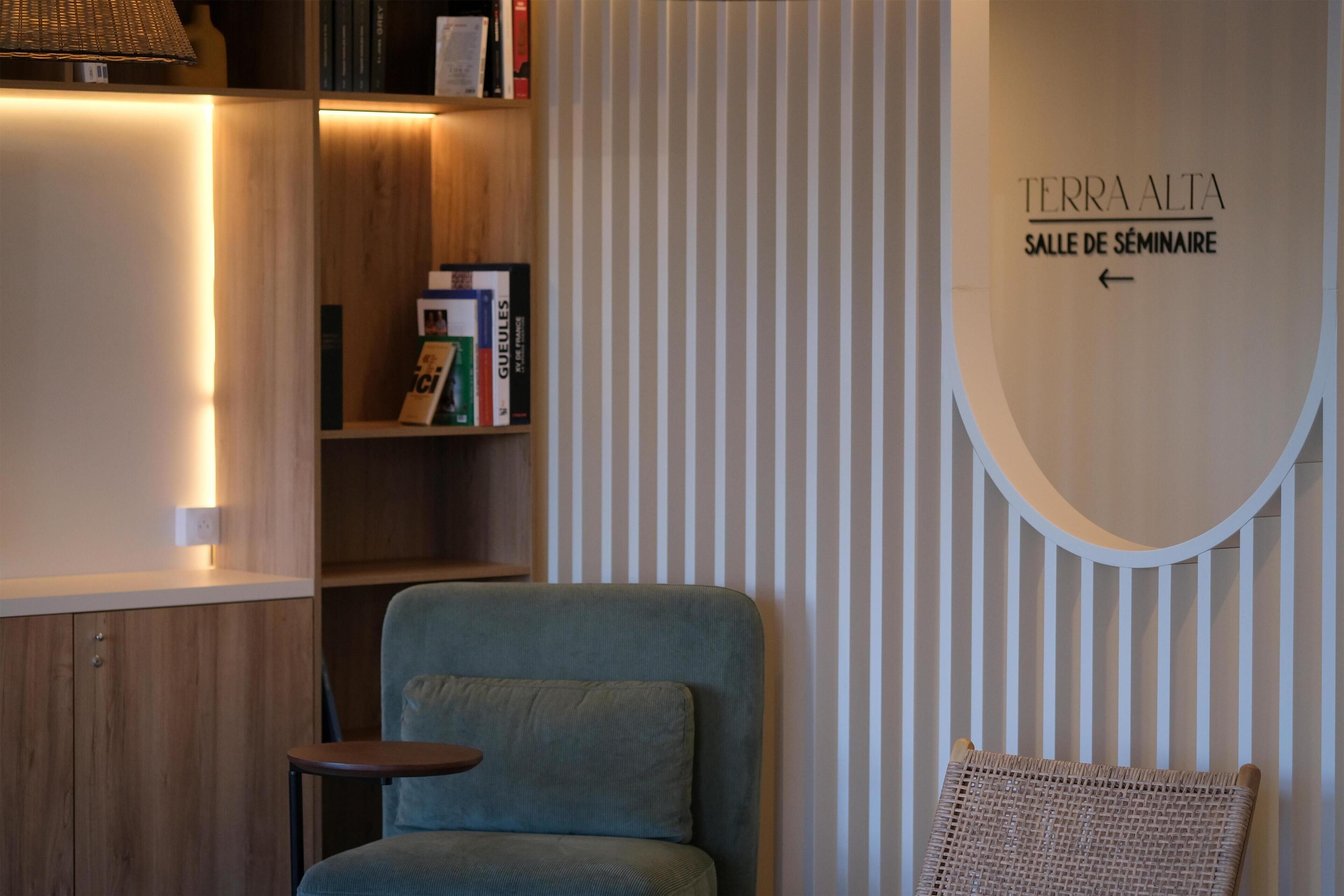 Une pièce moderne avec un fauteuil bleu, une petite table d’appoint, une bibliothèque et un panneau indiquant « TERRA ALTA, SALLE DE SÉMINAIRE ».