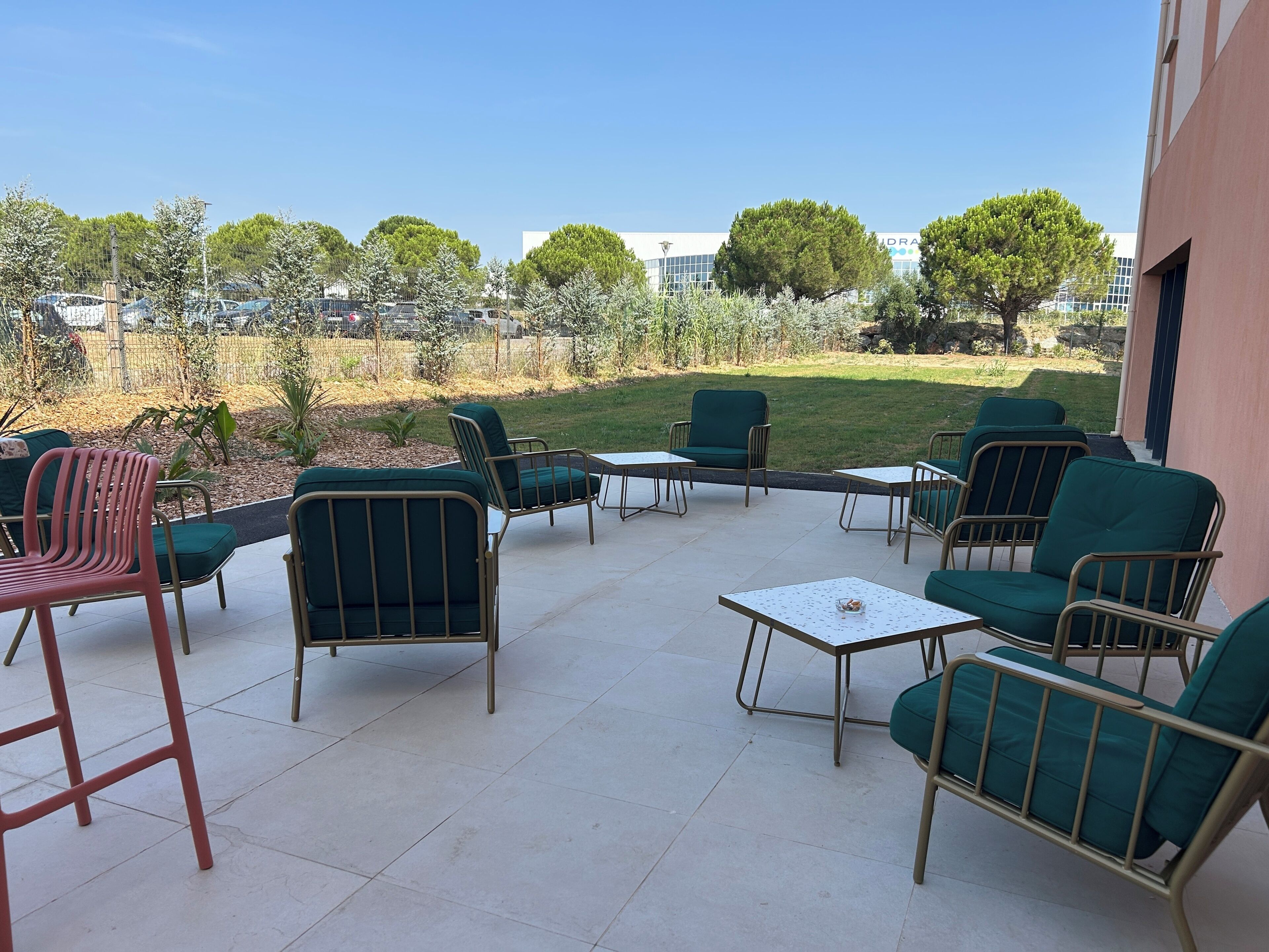 An outdoor patio with green chairs and tables, a grassy area, and a building in the background.