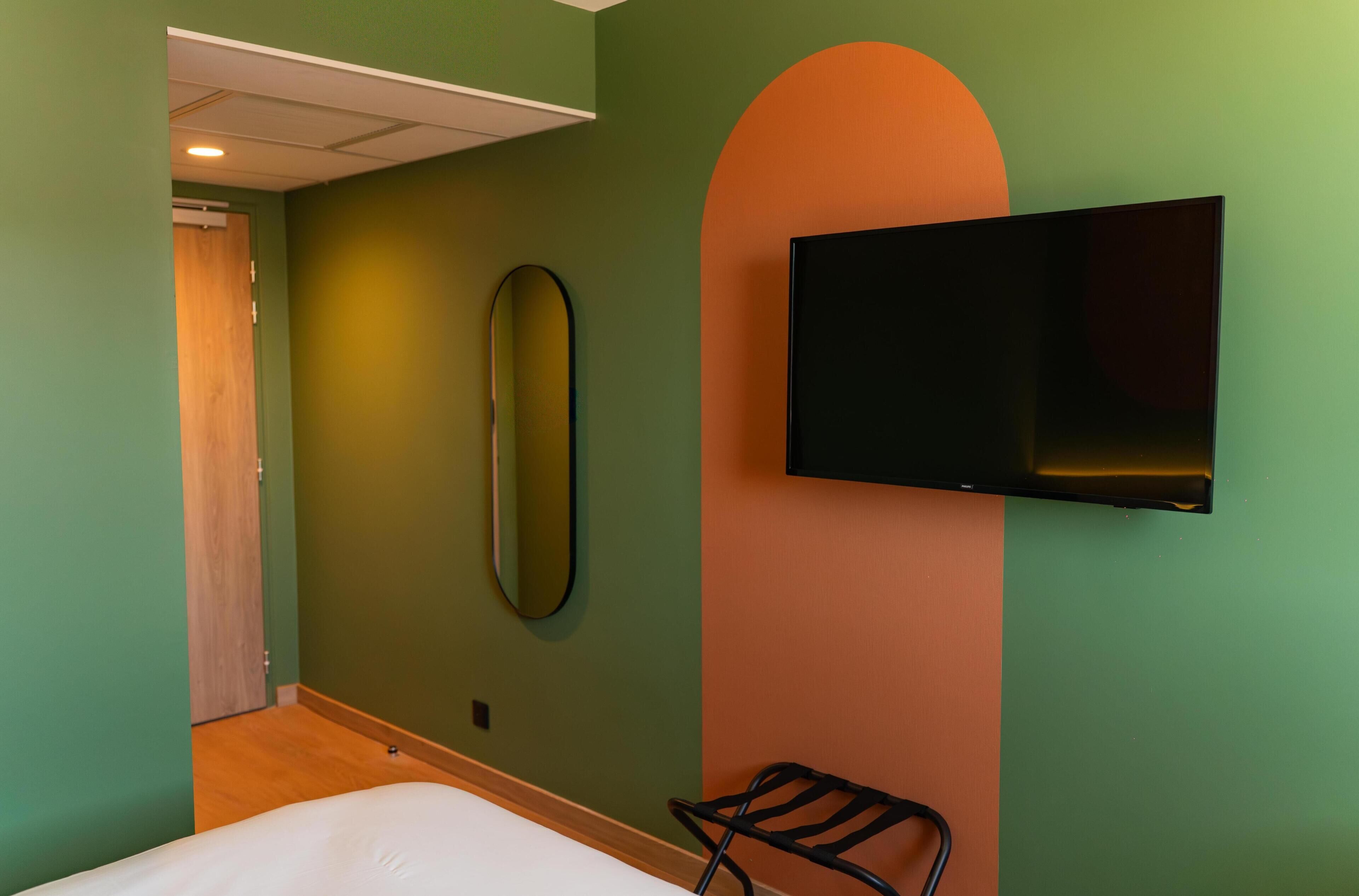 A room with a bed, a TV mounted on the wall, a mirror, and a door leading to another room.