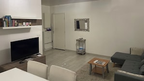 A living room with a flat-screen TV, a sofa, a coffee table, a bookshelf, and a door leading to another room.
