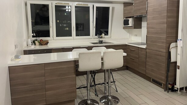 A modern kitchen with a white countertop, two bar stools, a window with frosted glass, and wooden cabinets.