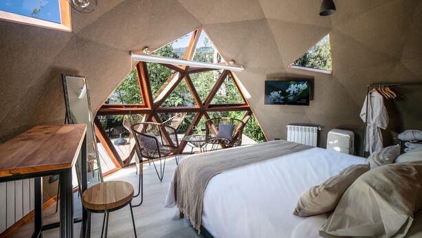 A bedroom with a large bed, a desk, and chairs, with a view of trees through triangular windows.