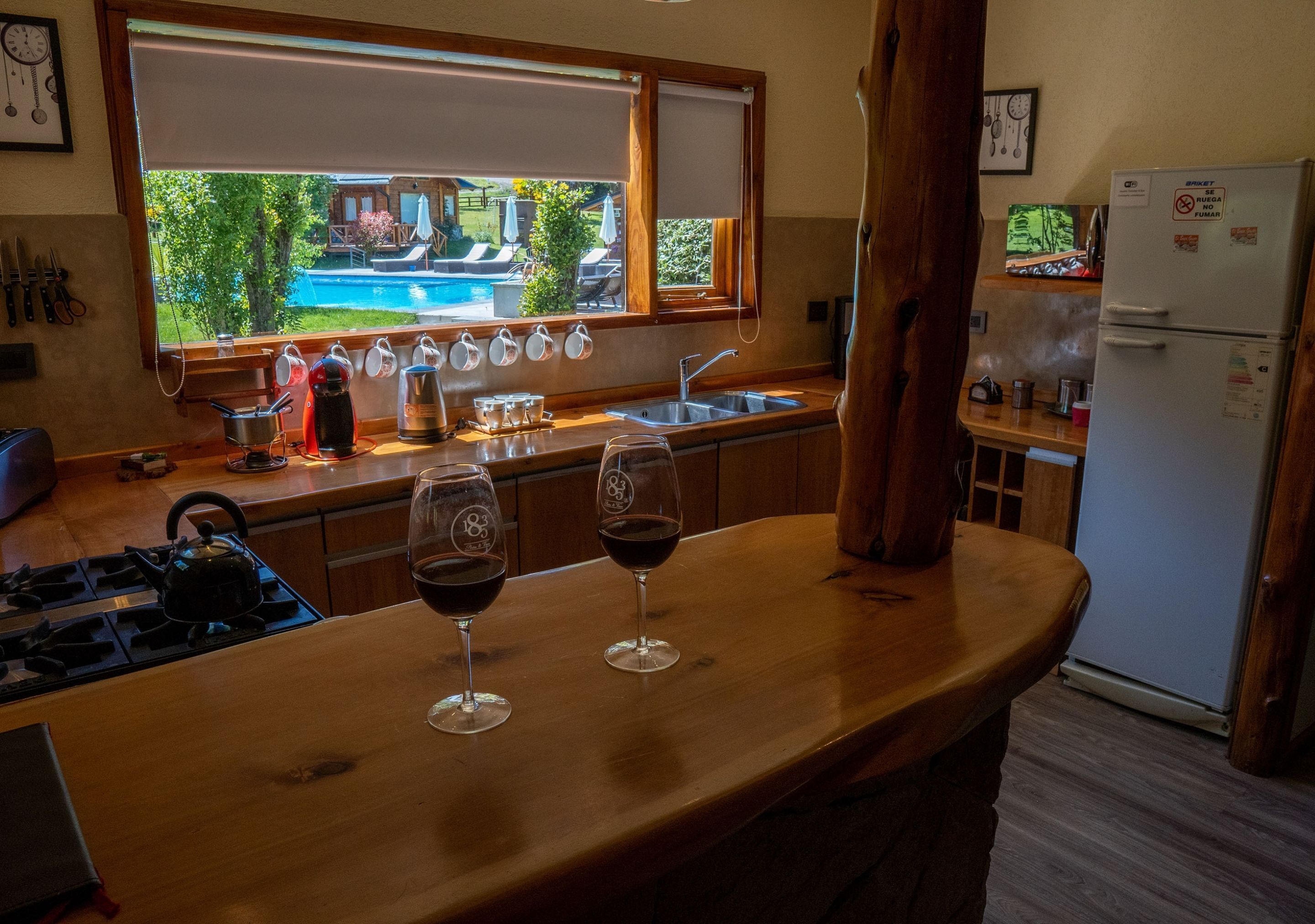 Cozinha com bancada de madeira, dois copos de vinho, chaleira e vista para uma piscina.