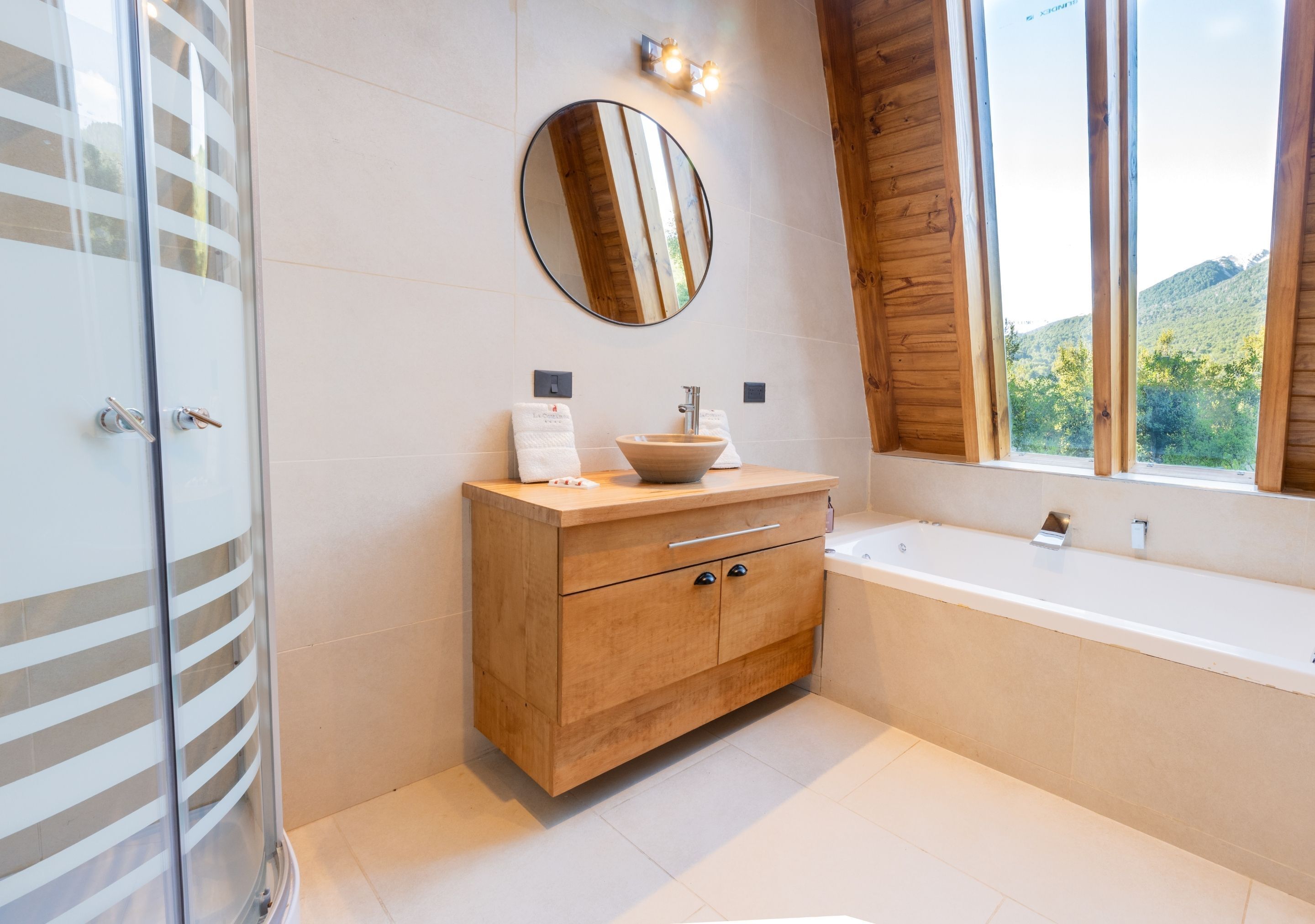 Un baño moderno con tocador de madera, un espejo redondo, una bañera y una ventana con vistas a la vegetación.